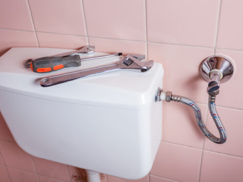 A toilet tank against a pink tiled wall with a wrench and screwdriver on top, next to a metal braided water supply line connected to a shut-off valve—different plungers like a sink plunger or toilet plunger aren't pictured but are helpful for maintenance. | High 5 Plumbing, Heating & Cooling