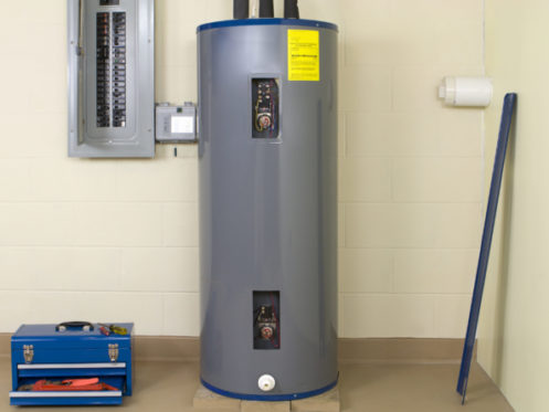 A gray traditional water heater stands on a concrete floor near a beige wall. To its left is an electrical panel, a blue toolbox, and some tools. A long blue strip leans against the wall, perfect for a water heater comparison. | High 5 Plumbing, Heating & Cooling