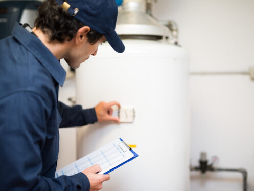 A person in a blue uniform and cap inspects a white boiler or water heater, holding a clipboard and adjusting the thermostat—a common scene for homeowners considering water heater replacement in their utility room. | High 5 Plumbing, Heating & Cooling