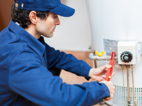 A person wearing a blue uniform and cap uses a red pipe wrench to adjust a component on a white water heater, showcasing emergency plumbing skills. Various pipes and a control panel are visible on the heater. | High 5 Plumbing, Heating & Cooling